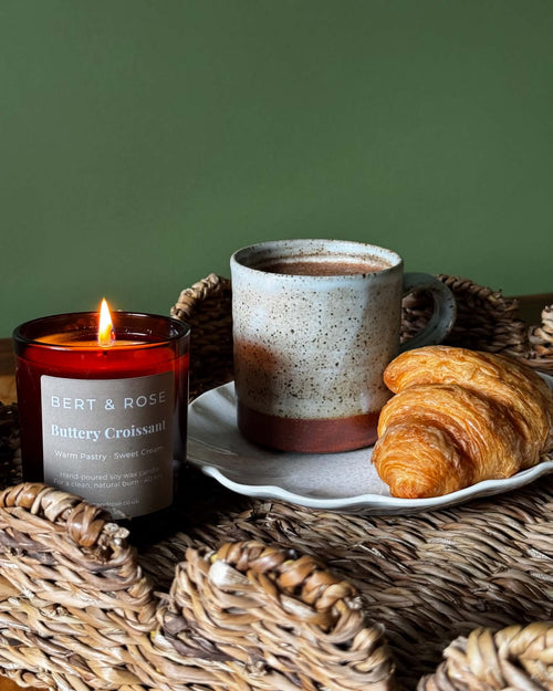 A lit amber glass candle labeled BERT & ROSE 'Buttery Croissant' sits on a woven tray beside a ceramic mug and a croissant on a white plate, against a muted green background.