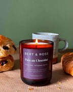 A lit candle in a dark amber glass jar with a purple label reading 'BERT & ROSE Pain au Chocolat' sits on a table with pastries and a ceramic mug in the background, against a muted green wall.