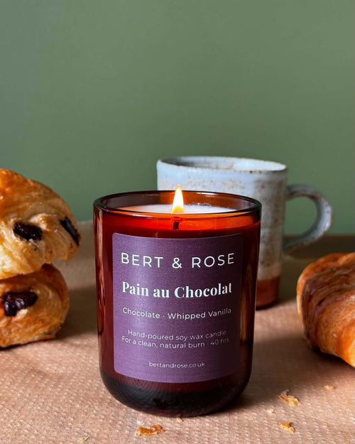 A lit candle in a dark amber glass jar with a purple label reading 'BERT & ROSE Pain au Chocolat' sits on a table with pastries and a ceramic mug in the background, against a muted green wall.