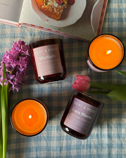 Three amber glass scented candles from Bert & Rose are arranged on a blue-and-white checkered tablecloth with flowers and an open book.