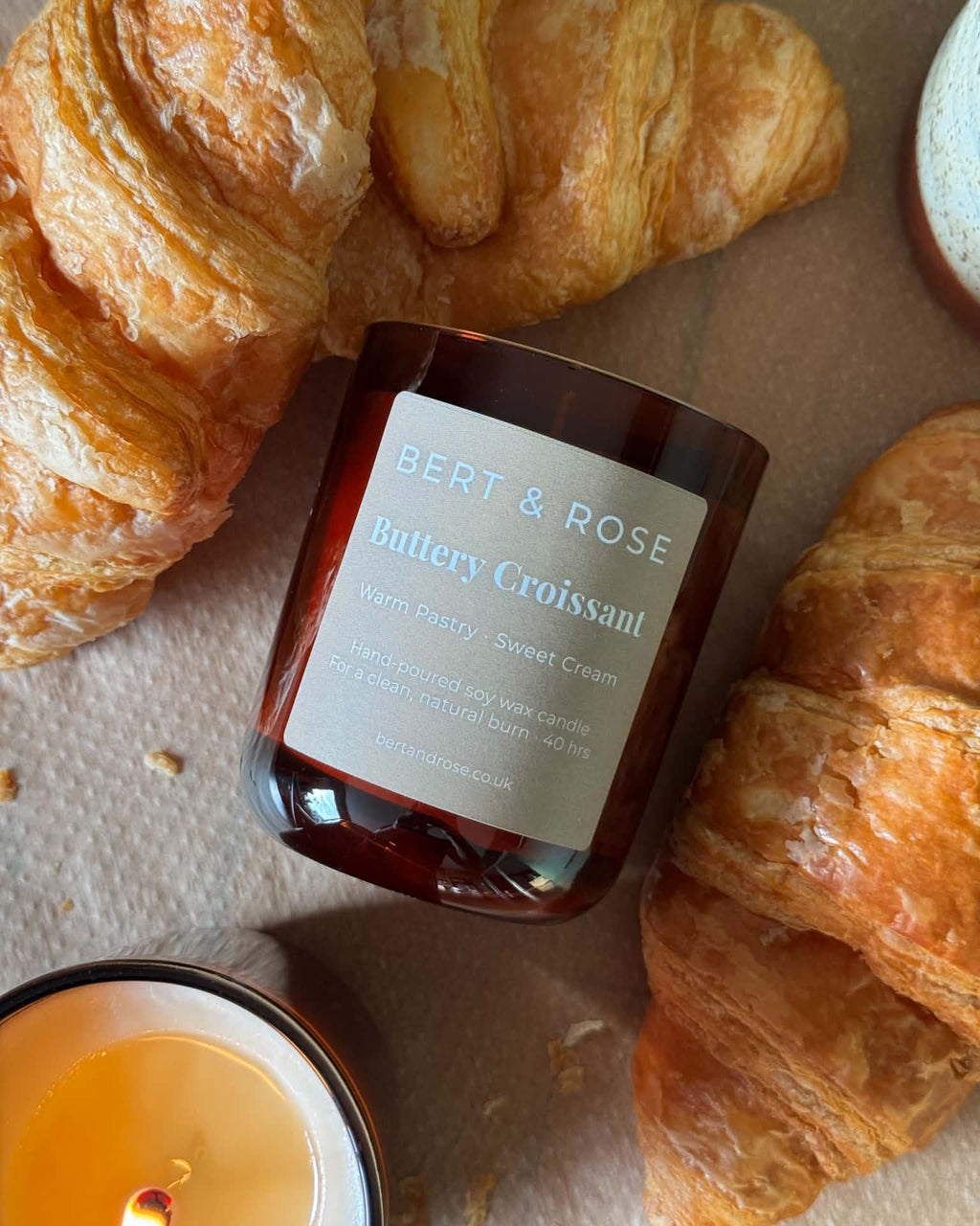 A Bert & Rose 'Buttery Croissant' scented candle in an amber glass jar, styled on a beige surface with fresh croissants and a lit candle nearby.