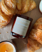 A Bert & Rose 'Buttery Croissant' scented candle in an amber glass jar, styled on a beige surface with fresh croissants and a lit candle nearby.