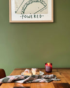 A framed art print with the text '-POWERED-' hangs on a sage green wall above a wooden dining table. On the table, a plaid napkin, a small plate with pastries, and a lit red candle are arranged.