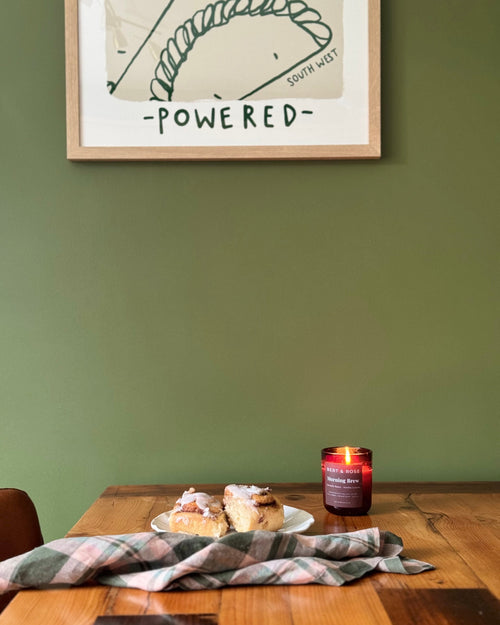 A framed art print with the text '-POWERED-' hangs on a sage green wall above a wooden dining table. On the table, a plaid napkin, a small plate with pastries, and a lit red candle are arranged.