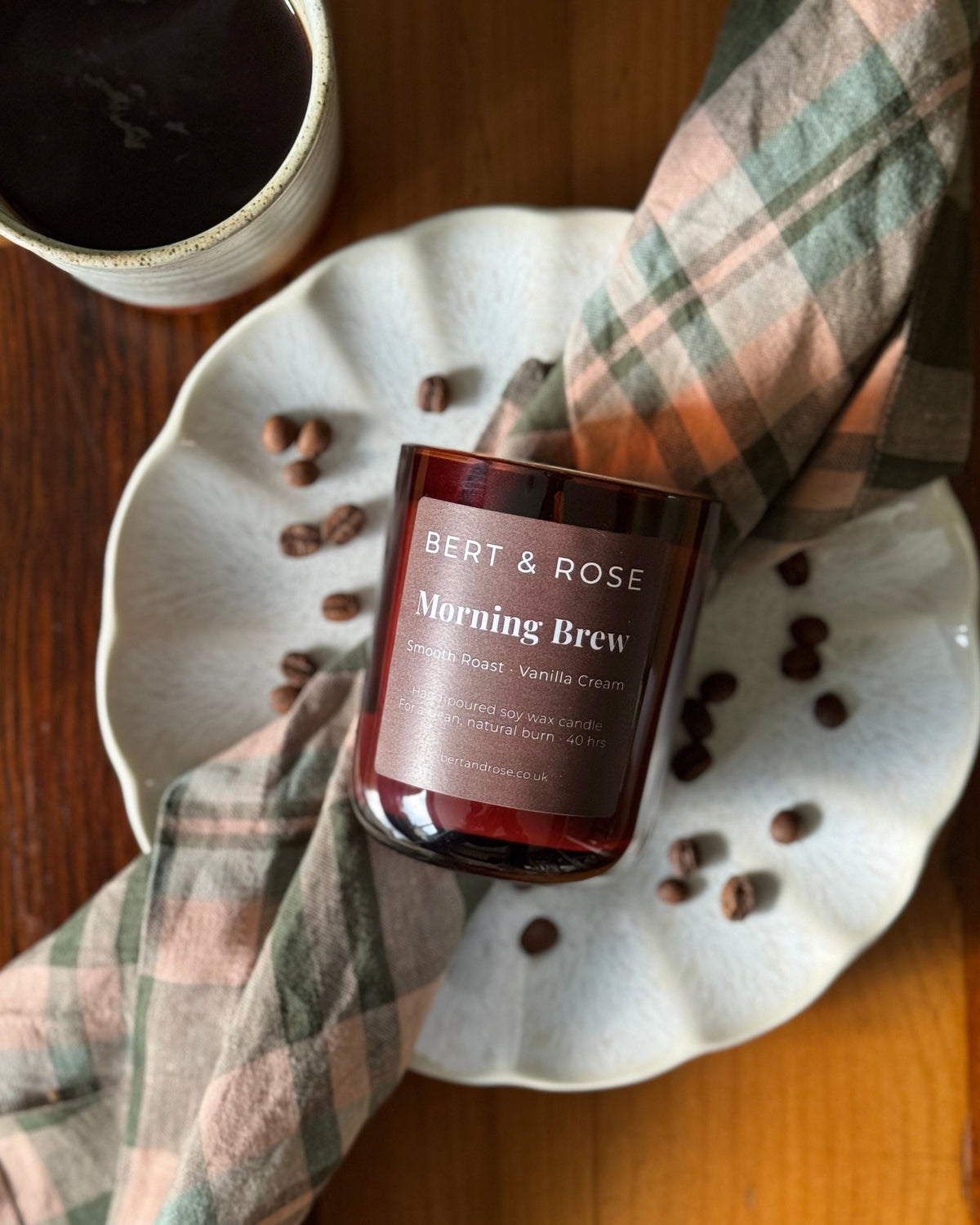 A jar candle labeled BERT & ROSE Morning Brew sits on a white scalloped plate with coffee beans scattered around it, styled with a plaid cloth on a wooden table next to a cup of coffee.