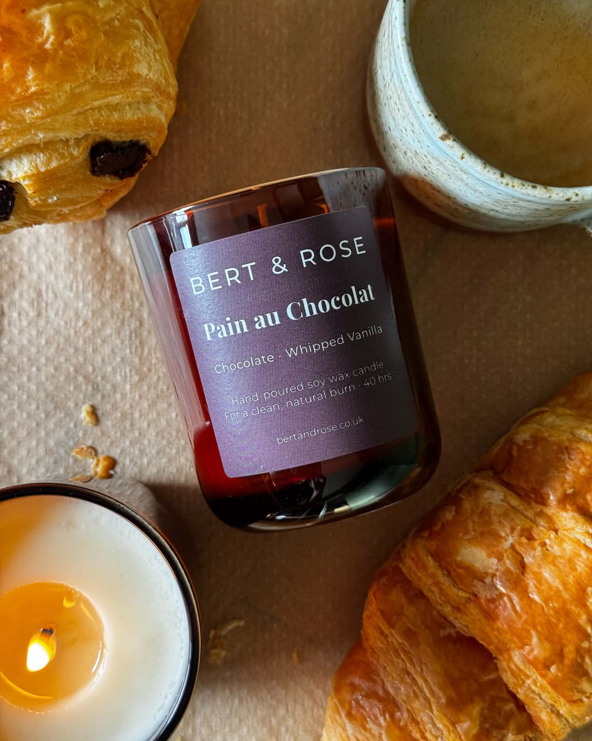 A jar candle labeled BERT & ROSE Pain au Chocolat, styled on a beige surface with croissants, a lit candle, and a ceramic cup.