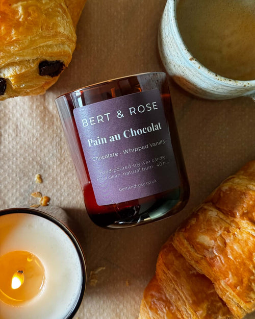 A jar candle labeled BERT & ROSE Pain au Chocolat, styled on a beige surface with croissants, a lit candle, and a ceramic cup.