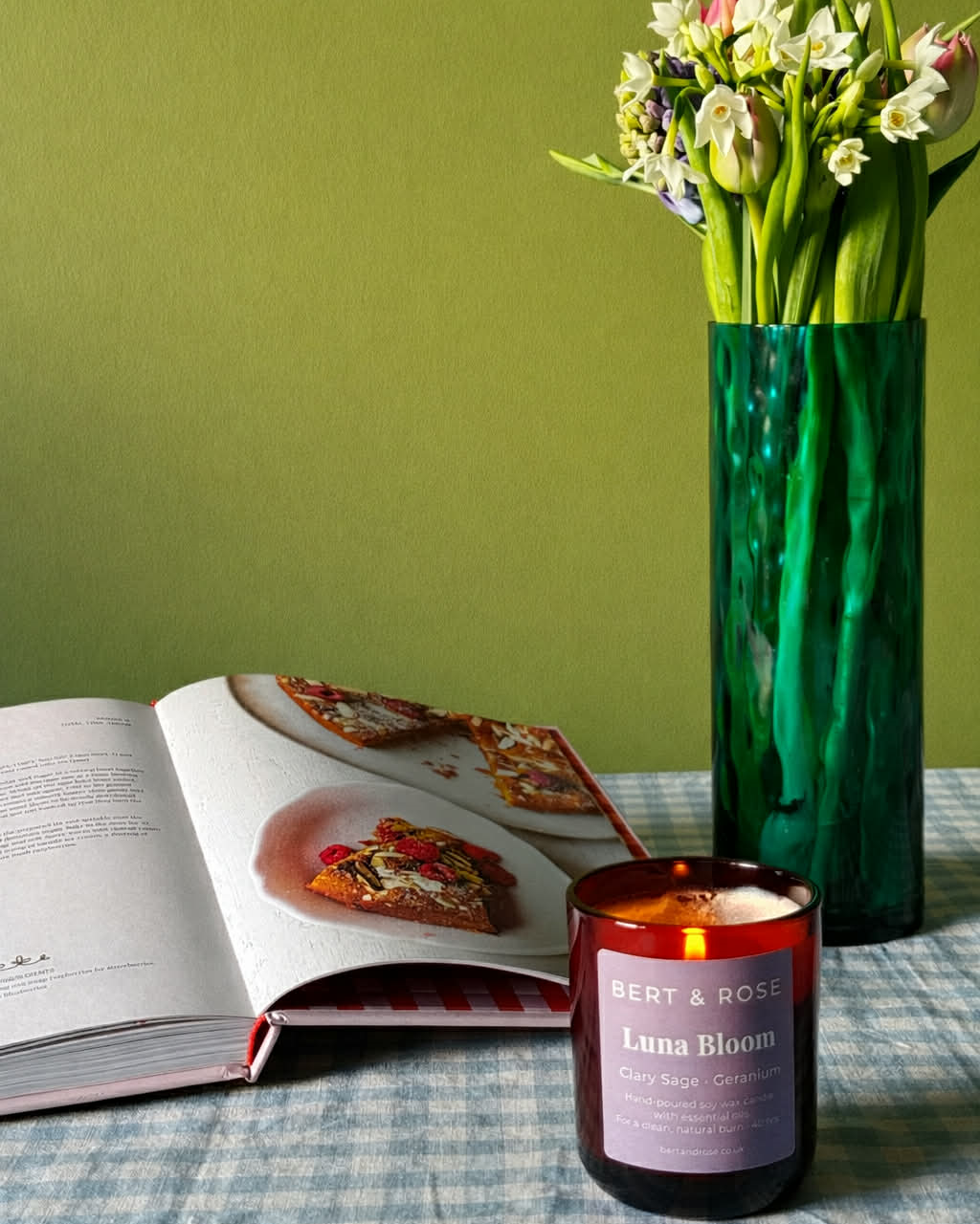 A lit amber glass candle jar labeled Bert & Rose Luna Bloom sits on a blue-and-white checkered tablecloth next to an open magazine and a tall green vase holding tulips against an olive green wall.