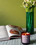 A lit amber glass candle jar labeled Bert & Rose Luna Bloom sits on a blue-and-white checkered tablecloth next to an open magazine and a tall green vase holding tulips against an olive green wall.